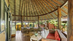 Een overdekt terras met een tafel, stoelen, een rieten bank en een plant in pot.
