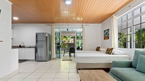 Una habitación moderna con cama, un sofá verde, techo de madera, zona de cocina y vistas al verdor.