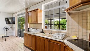 A kitchen with wooden cabinets, a double sink, a fan, and a large window with a view of greenery. - Moorea Mahana House With Private Canal to Lagoon (Moorea-Maiao)