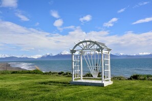 A white gazebo with a circular roof, located on a grassy area with a view of a lake and snow-capped mountains in the background. - Stunning Beachside Vacation Rental for Family or Group Glamping Escapes to Alaska (Homer)