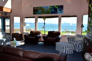 A room with a large window overlooking a body of water, leather furniture, and a painting above the window. - Stunning Beachside Vacation Rental for Family or Group Glamping Escapes to Alaska (Homer)