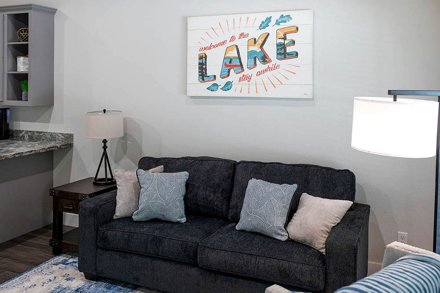 A living room with a dark grey sofa, two decorative pillows, a framed welcome sign, a floor lamp, and a kitchen area with a cabinet and countertop.