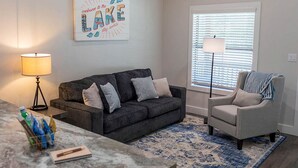 Una sala de estar con un sofá gris oscuro, un sillón gris claro, un tapete estampado azul, una lámpara y un letrero que dice 'welcome to the LAKE stay awhile'.
