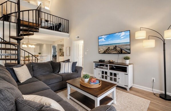 Ein modernes Wohnzimmer mit einem grauen Ecksofa, einem Holztisch, einem an der Wand montierten Fernseher, einer Treppe und einem weißen Schrank.