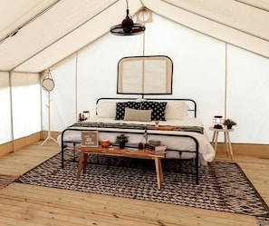 A tented room with a bed, a small table, a hanging light fixture, and a side table with a plant.