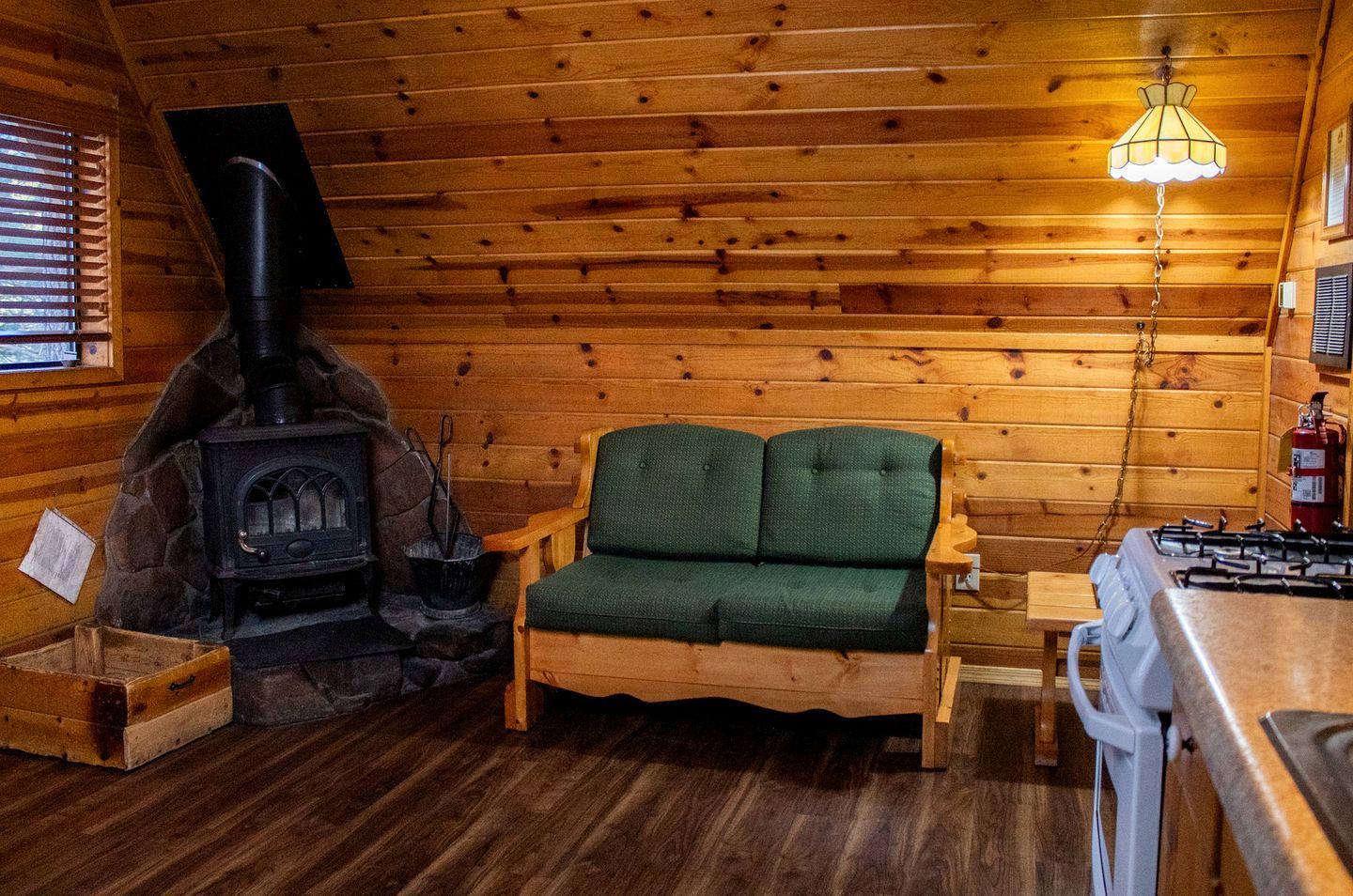 Interior acogedor de una cabaña con chimenea de piedra, sofá tapizado en verde, piso de madera y una zona de cocina con estufa a gas.