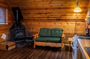 L’intérieur chaleureux d’un chalet avec une cheminée en pierre, un canapé recouvert d’un tissu vert, un plancher en bois et un coin cuisine équipé d’une cuisinière à gaz.