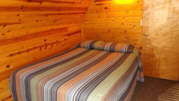 Une pièce chaleureuse avec un plafond en bois à pente, un lit recouvert d’une courtepointe à rayures et une porte surmontée d’un luminaire.