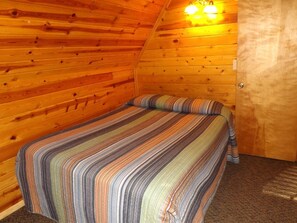 A cozy room with a wooden sloped ceiling, a bed with a striped bedspread, and a door with a light fixture above it. - Charming Equipped Cabin Suitable for Four in Arizona (Flagstaff)