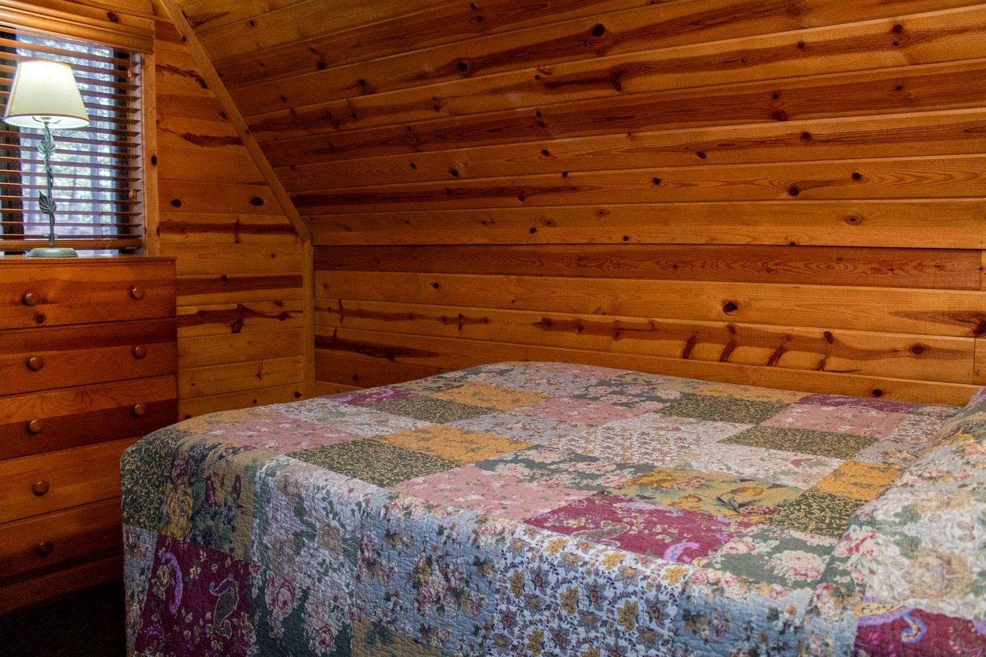 Een slaapkamer met een houten plafond, een bed met een bloemenquilt, een houten dressoir en een raam met jaloezieën.