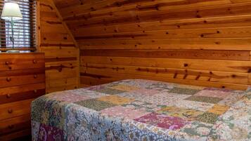 Een slaapkamer met een houten plafond, een bed met een bloemenquilt, een houten dressoir en een raam met jaloezieën.