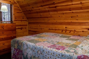 Une chambre avec un plafond en bois, un lit recouvert d’une courtepointe à motifs floraux, une commode en bois et une fenêtre avec des stores.