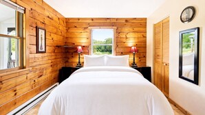 A bedroom with a wooden paneled wall, a bed with white bedding, two bedside lamps, a window with a view of greenery, and a clock on the wall. - Luxury Catskills Creekside Cabin | Pool, Sauna & Fire Pit Retreat (Roscoe)