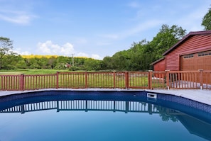 Une piscine dotée d’une terrasse en bois et d’une clôture, entourée de verdure et bordée d’un bâtiment rouge.