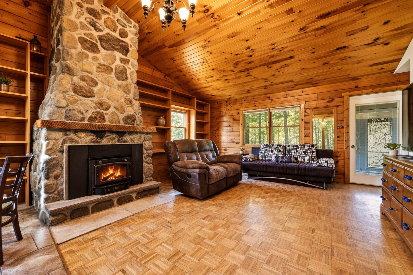 A log cabin interior with a stone fireplace, wooden floors, a leather sofa, and a large window.