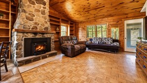 A log cabin interior with a stone fireplace, wooden floors, a leather sofa, and a large window. - Luxury Catskills Creekside Cabin | Pool, Sauna & Fire Pit Retreat (Roscoe)