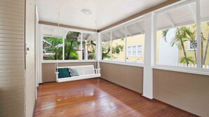 Ein überdachter Balkon mit Holzboden, einer weißen Schaukel und großen Fenstern mit Blick auf Palmen und ein gelbes Gebäude.