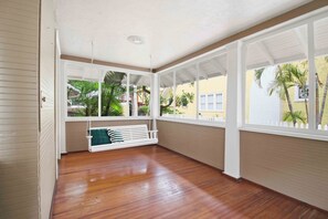 A covered porch with a wooden floor, a white swing, and large windows with a view of palm trees and a yellow building. - The Veranda at Wares Creek - Sleeps 12, Pet-friendly, minutes to Gulf beaches! (Bradenton)