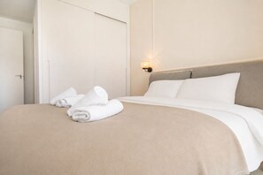 Ein ordentlich bezogenes Bett mit weißen Laken und einer beigen Tagesdecke, eine wandmontierte Leuchte und eine geschlossene Tür.