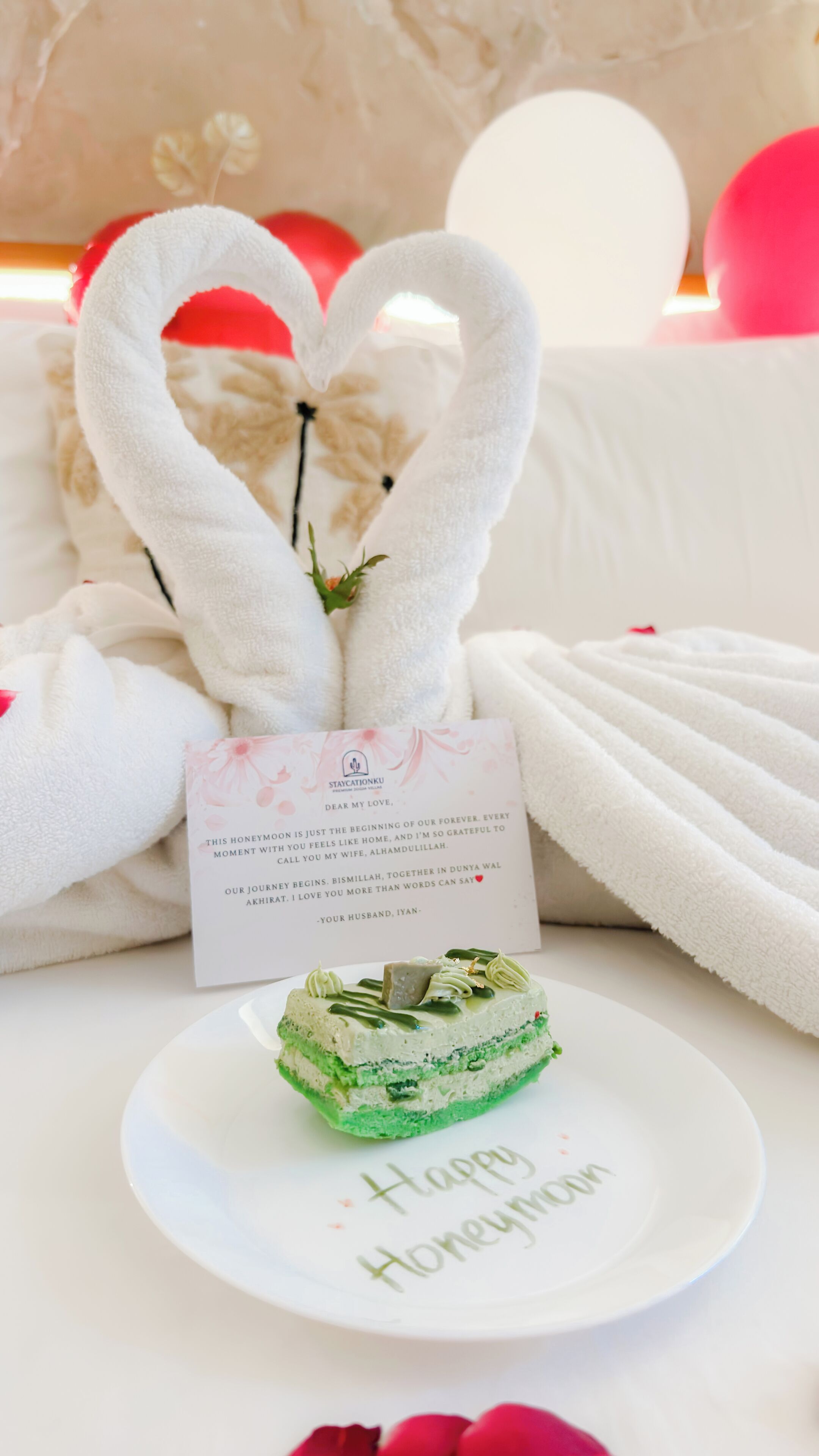 A wedding cake on a plate with 'Happy Honeymoon' written on it, surrounded by white towels and a card with a message.