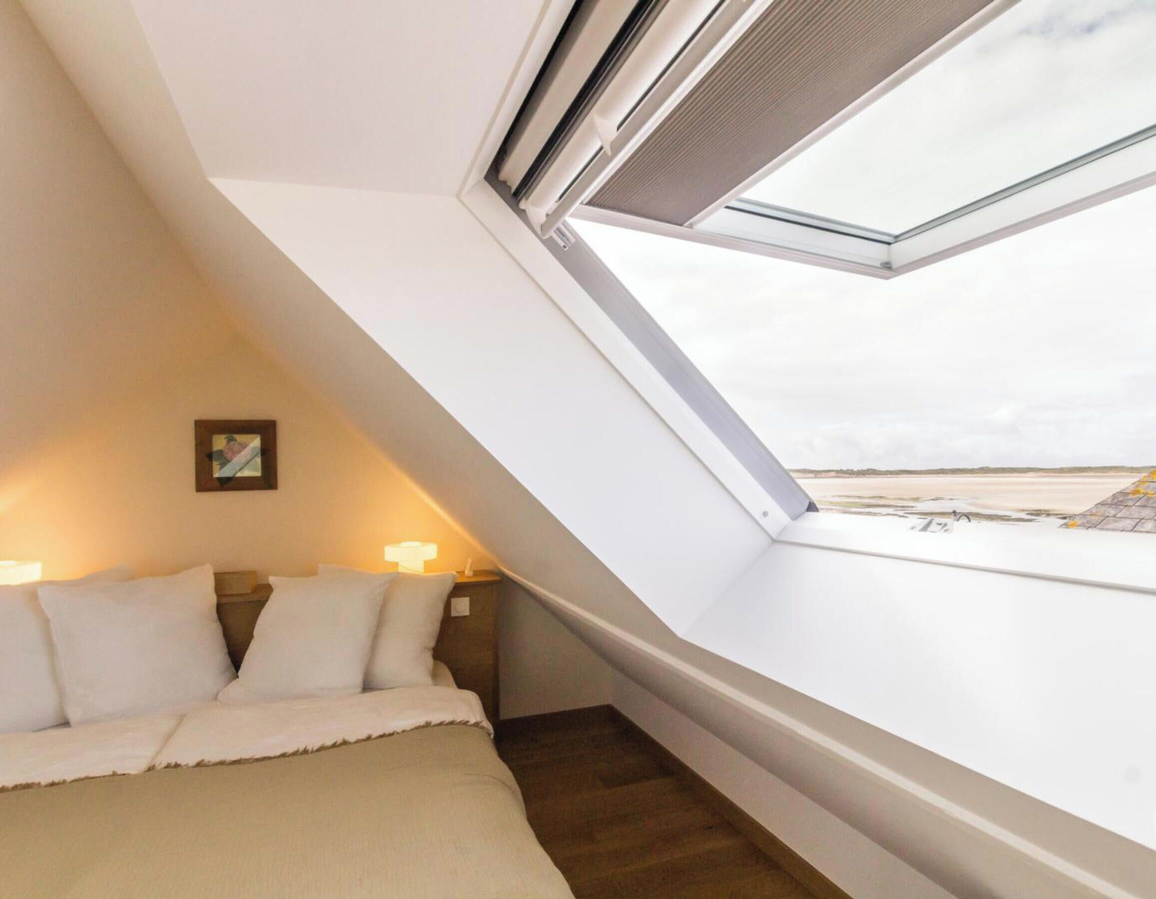 A bedroom with a sloped ceiling, a bed, a nightstand, a lamp, and a window overlooking a beach.