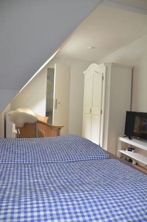 A bedroom with a checkered bedspread, a white wardrobe, a television on a stand, and a chair with a white cushion.