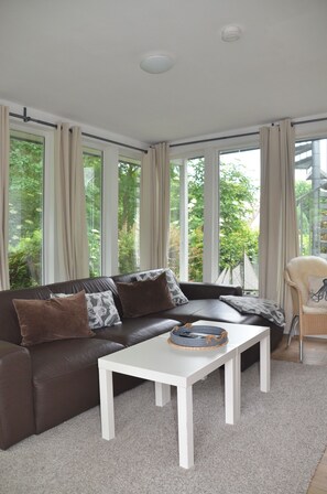 Un salon moderne avec un canapé en cuir marron, une table basse blanche et une vue sur la verdure grâce à de grandes fenêtres.