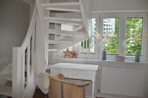 Un escalier blanc offre une vue sur la verdure à l’extérieur. On y trouve un radiateur blanc, une chaise en osier et des étoiles décoratives posées sur une étagère.