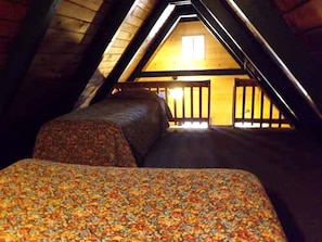 Une chambre mansardée avec un plafond en pente, deux lits avec une literie à motifs floraux et une vue sur une structure en bois à l’extérieur.