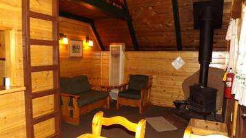 L’intérieur chaleureux d’un chalet avec des murs en bois, une cheminée, deux fauteuils verts rembourrés, une table en bois et une échelle menant à un niveau supérieur.