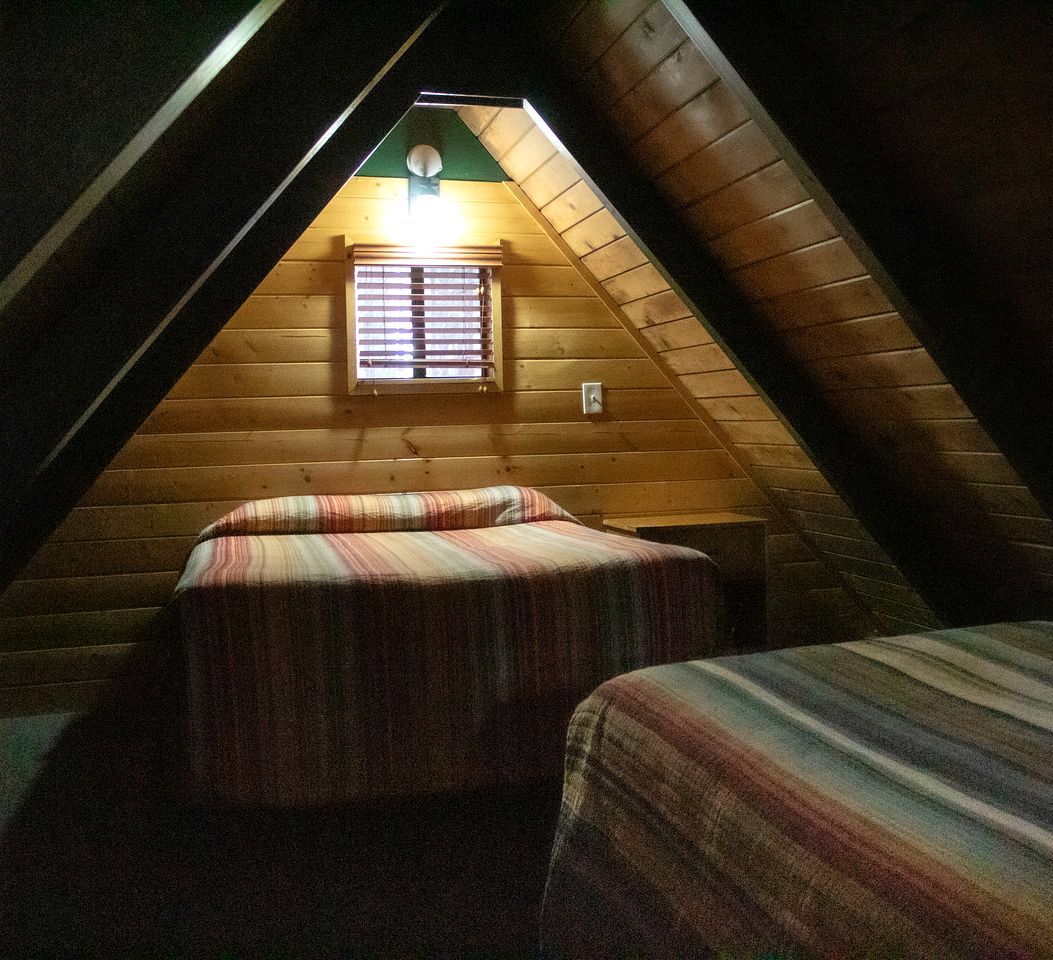 A small, cozy attic bedroom with two beds, a sloped ceiling, and a window with blinds.