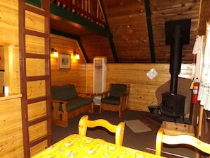 L’intérieur chaleureux d’un chalet avec des murs en bois, une cheminée, deux fauteuils verts rembourrés, une table en bois et une échelle menant à un niveau supérieur.