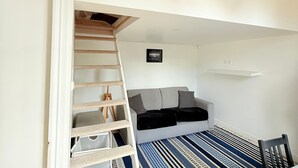 Una habitación con cama tipo litera, un sofá pequeño, una escalera, un cuadro enmarcado y una estantería.