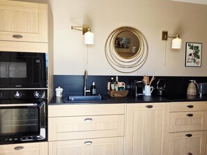 A modern kitchen with a built-in oven, a microwave, a sink with a faucet, and a coffee maker. There are wooden cabinets and a black countertop.
