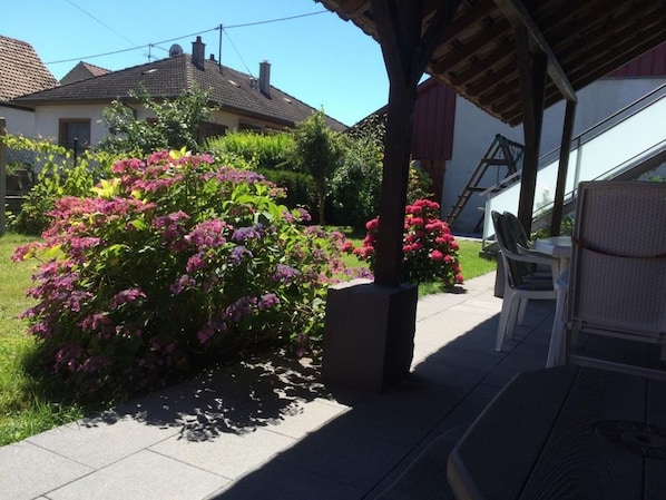 A covered patio area with a table and chairs, surrounded by lush greenery and flowering bushes.