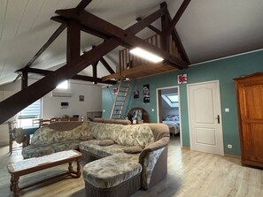 A loft-style room with exposed wooden beams, a large sectional sofa, a wooden bench, and a ladder leading to a lofted area.