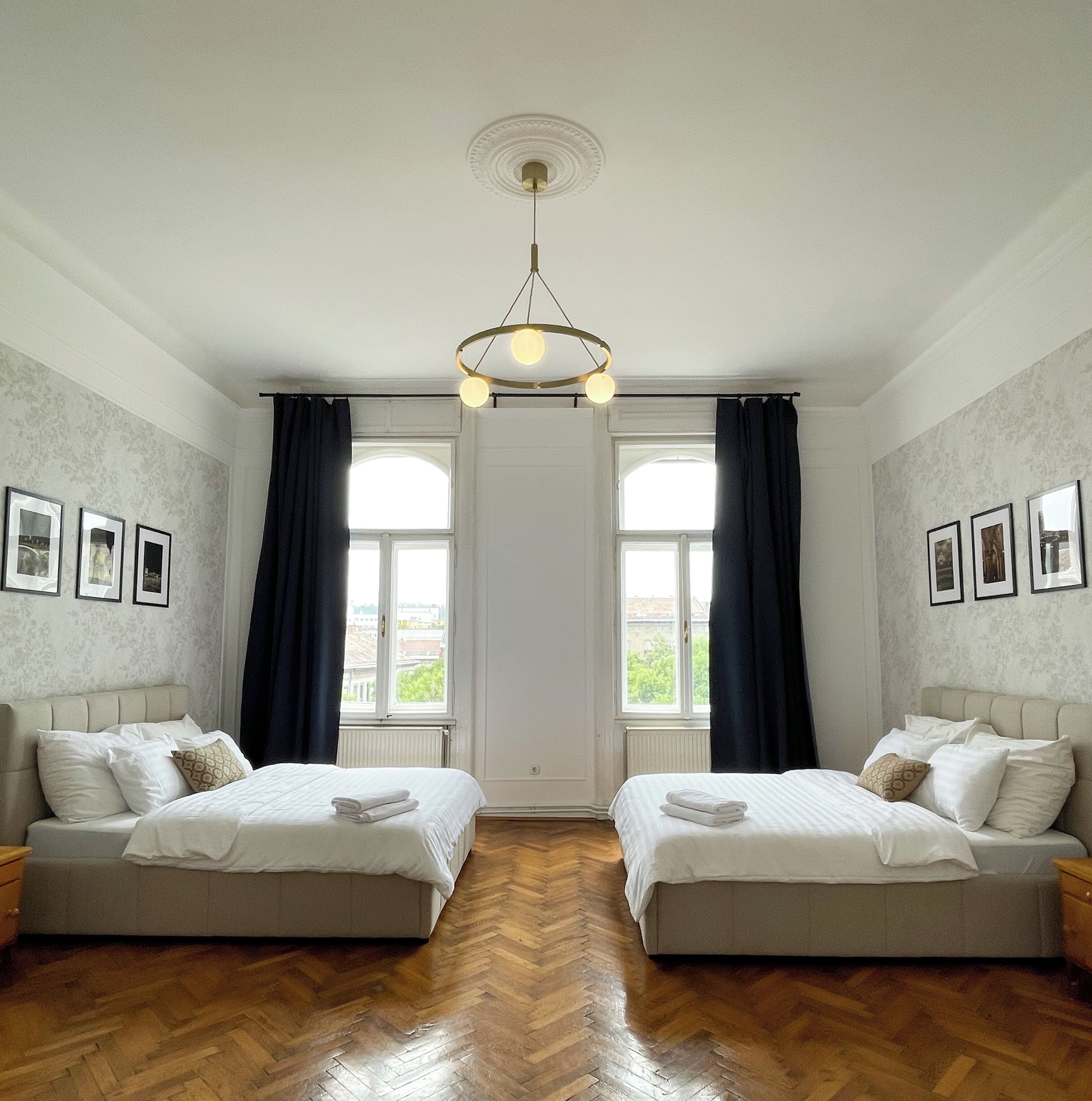 Quarto de hotel com duas camas, piso de madeira em padrão herringbone, luminária no teto e quadros nas paredes.
