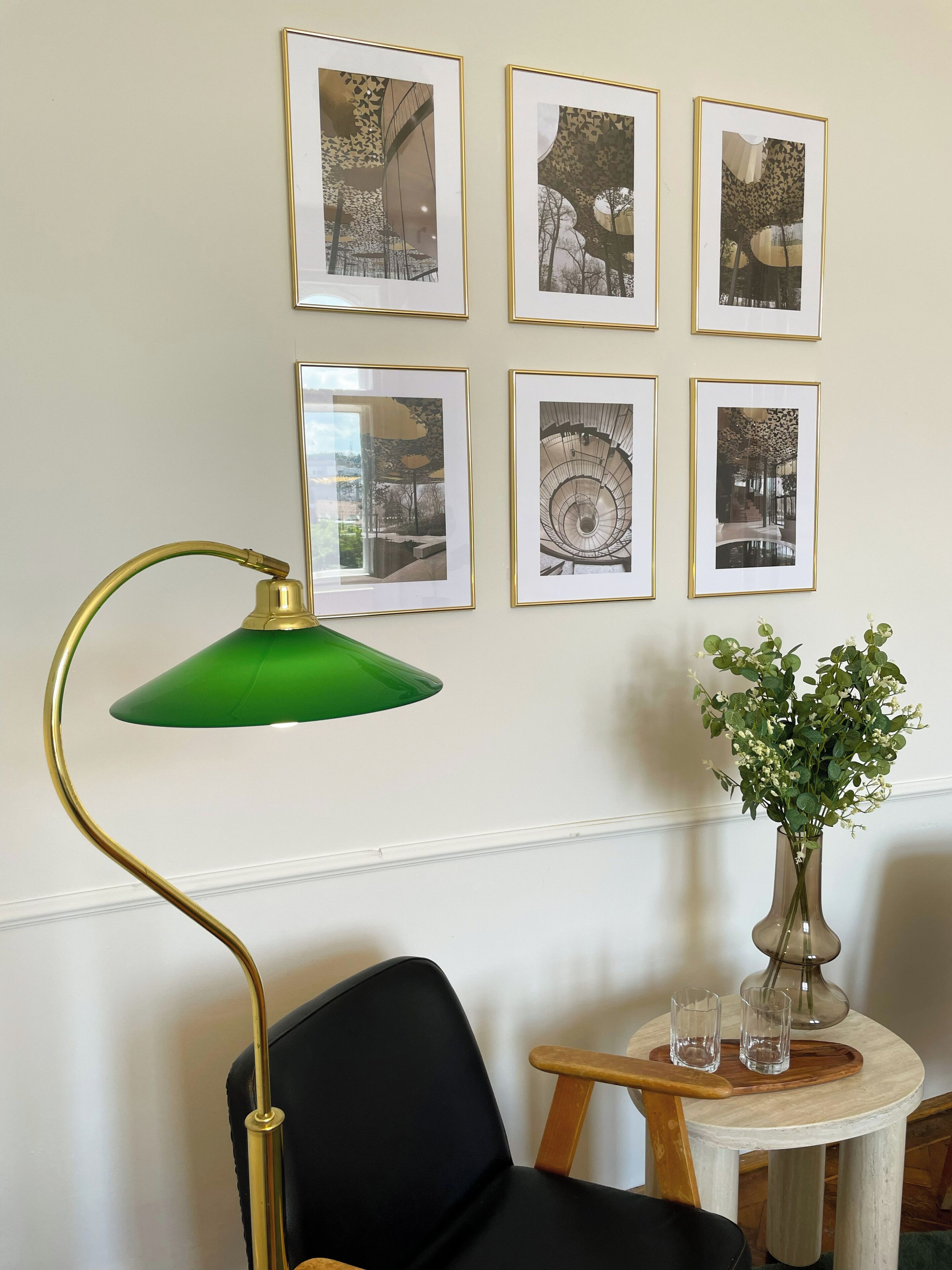 Una habitación con una silla negra, una lámpara de pie verde, una pequeña mesa redonda con un florero y seis cuadros en la pared.