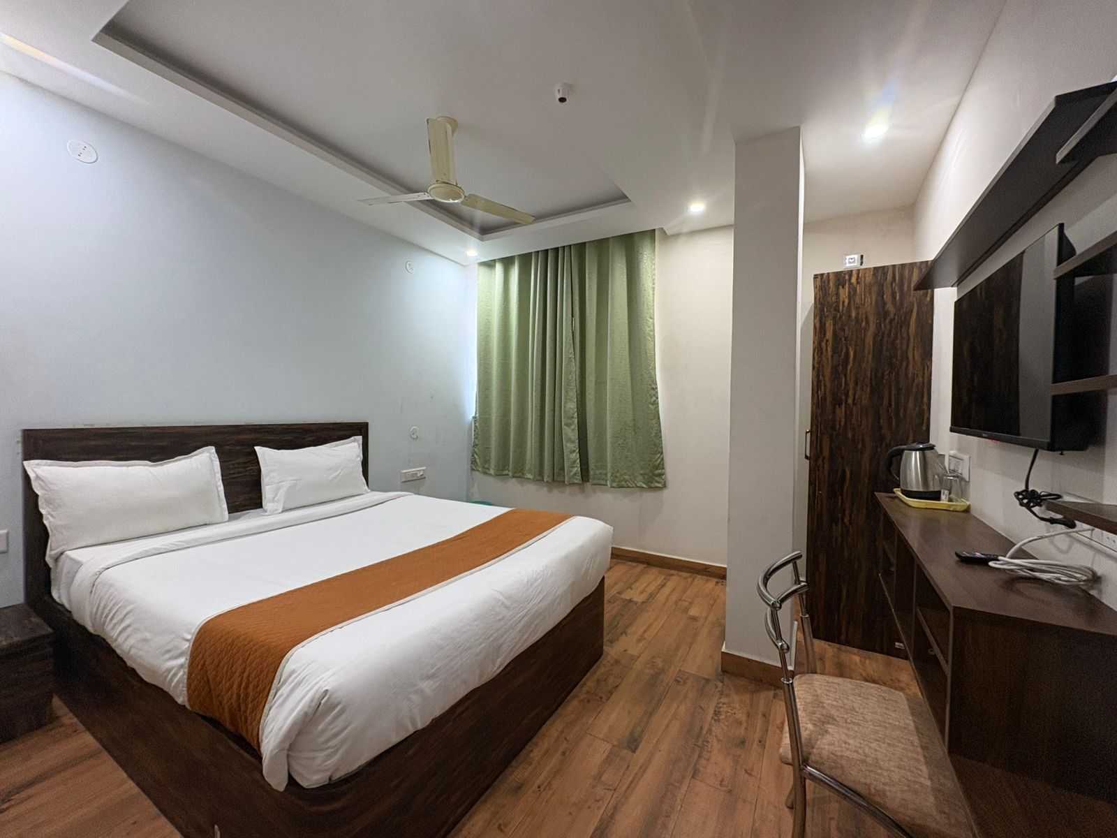 A hotel room with a bed, a desk with a TV, a chair, a window with curtains, and a ceiling fan.