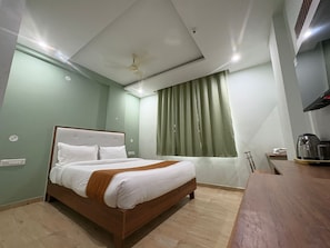 A hotel room with a bed, a ceiling fan, a window with curtains, a coffee machine, and a wooden desk. - Hotel Raasaa Grand Rooms (Bengaluru)