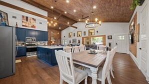 A spacious kitchen with wooden cabinets, a large island, and a dining area with a wooden table and white chairs. - Terracotta Retreat · Sleeps 54 · Pool & Hot Tub (Wimberley)