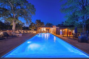 En pool med ett uteområde, en möblerad plats under tak och en byggnad med varm belysning.