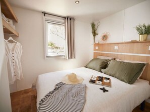 A bedroom with a bed, a window, a shelf with plants, and a hanging garment. - Mobile home with AC - 4 guests (Arles-sur-Tech)