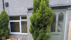 Um imóvel residencial com porta de entrada, um pequeno jardim com gramado e uma árvore, e um pátio com mesa e cadeiras.