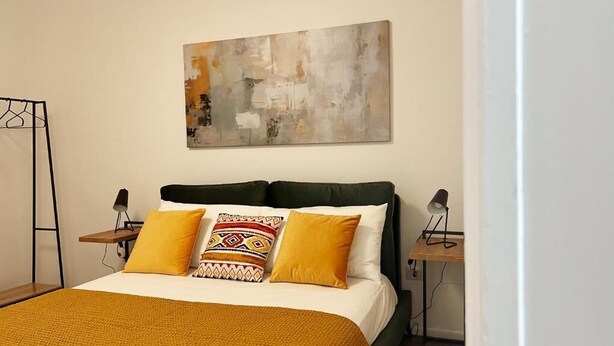 Una cama con una colcha blanca y mostaza, dos almohadas, un cabecero verde y un cuadro abstracto colgado en la pared.