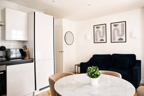 A modern kitchen with white cabinets, a black countertop, and a coffee machine. There is a round table with a potted plant in the living area with a dark blue sofa. - Charming 1BR with Balcony in Central Fitzrovia (London)
