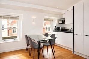 A compact kitchen with a dining table and chairs, a refrigerator, and a view of the street. - Fitzrovia and West End Residences (London)
