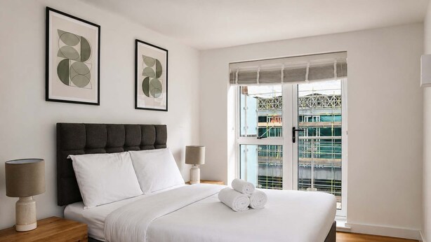 Ein Schlafzimmer mit einem Bett, zwei Nachttischen, einem Fenster mit Blick auf eine Baustelle und zwei gerahmten Kunstwerken an der Wand.