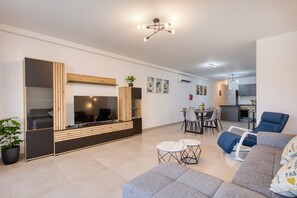 Ein modernes Wohnzimmer mit Sofa, Fernseher, Essbereich und einer Küche im Hintergrund.