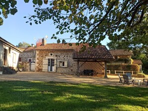 Un edificio rústico de piedra con techo de tejas, un porche cubierto con mesas al aire libre y un césped bien cuidado.